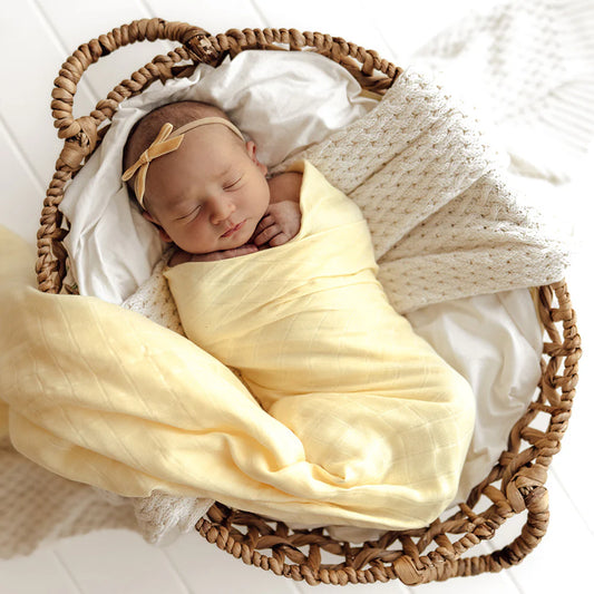 Newborn baby wrapped in a yellow blanket with a headband, lying in a wicker basket.