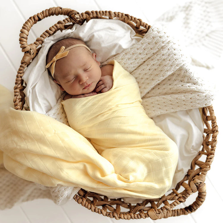 Newborn baby wrapped in a yellow blanket with a headband, lying in a wicker basket.
