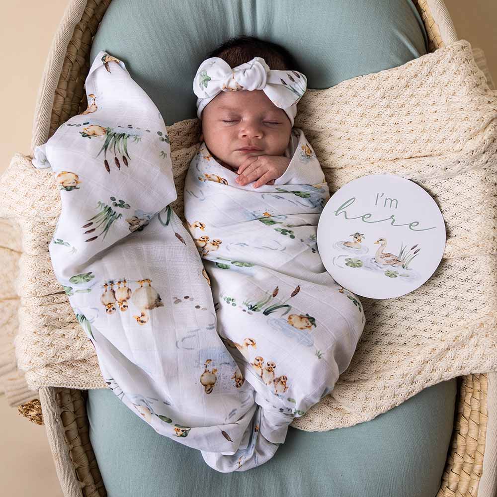 Newborn baby wrapped in a swaddle blanket with ducks on it and a 'I'm here' sign in a wicker basket.