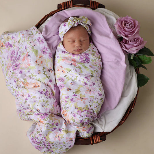 Newborn baby wrapped in a floral swaddle with a matching headband, surrounded by pink flowers in a wooden basket.