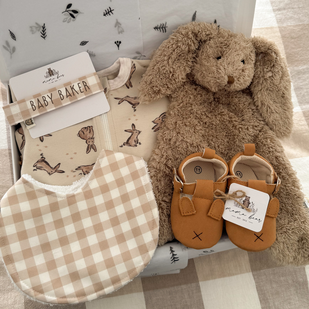 New baby gift box featuring beige tones. A snuggly light brown bunny rabbit toy comforter, tan bunny baby shoes, a beige and cream gingham bib, matching dummy clip personalised with babies name and a cream bodysuit with illustrations of brown bunnies scattered on it.