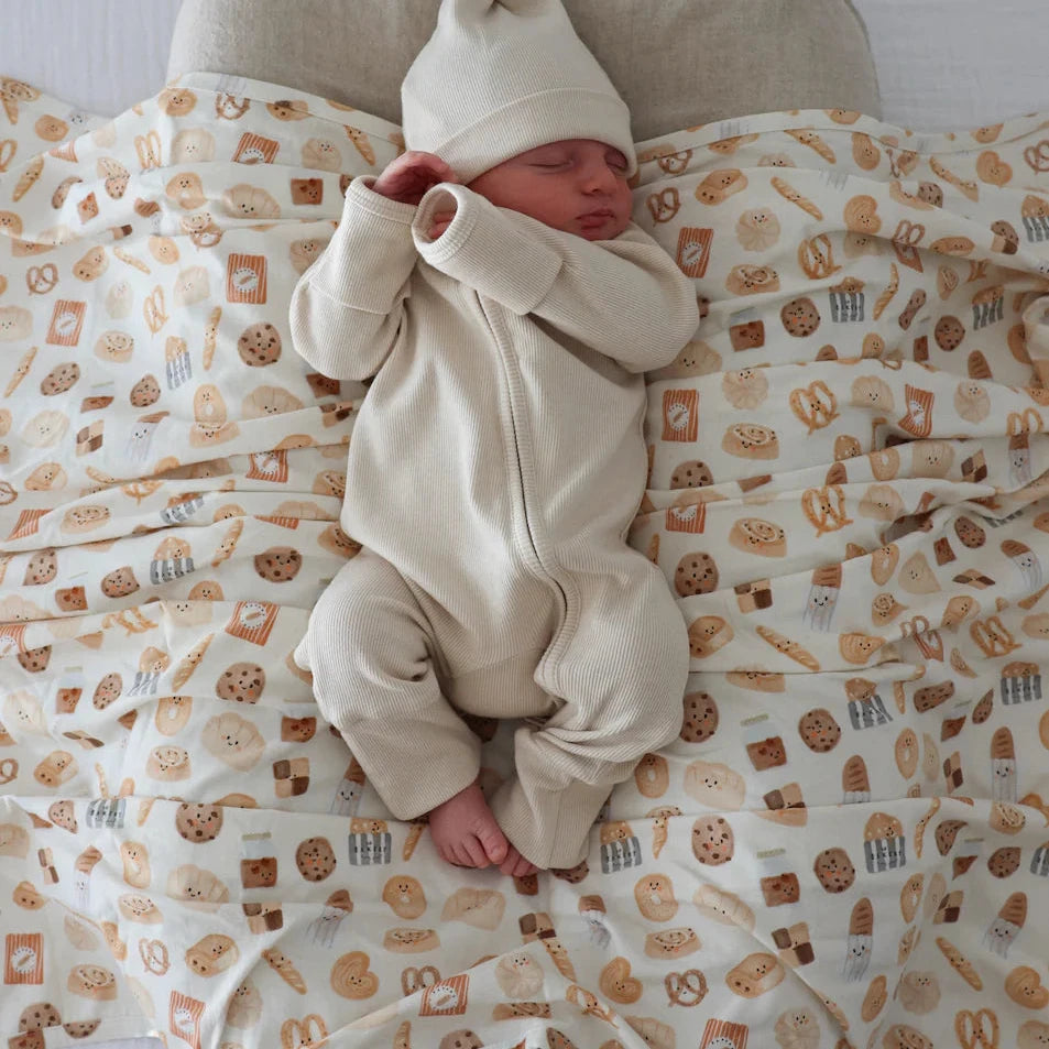Newborn baby in a white outfit and hat lying on a patterned bread swaddle.