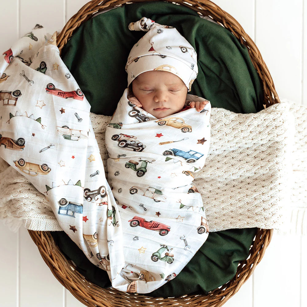 Newborn baby swaddled in a blanket with car pattern, wearing a matching beanie, in a wicker basket.