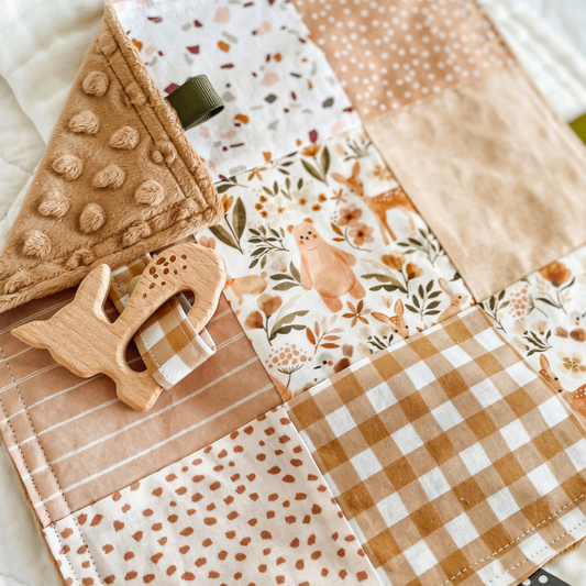 Handmade baby Taggie blanket in warm earthy tones with a mix of patchwork prints including gingham, florals, terrazzo, stripes, and woodland animals. Features a wooden deer-shaped teether attached with a matching fabric strap, textured minkee backing, and neutral-toned ribbon loops for sensory play.