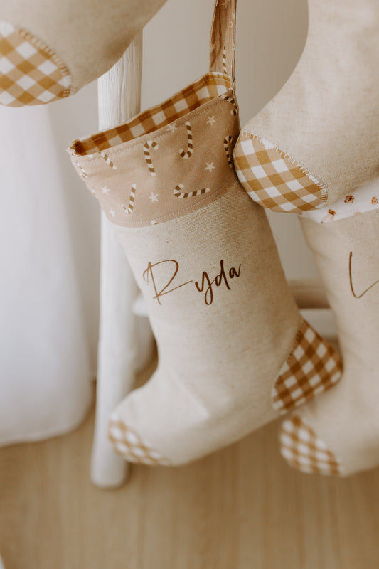 Handmade personalised Christmas stocking in beige fabric with caramel gingham heel and toe, featuring a candy cane and star print cuff, customised with name Ryda.