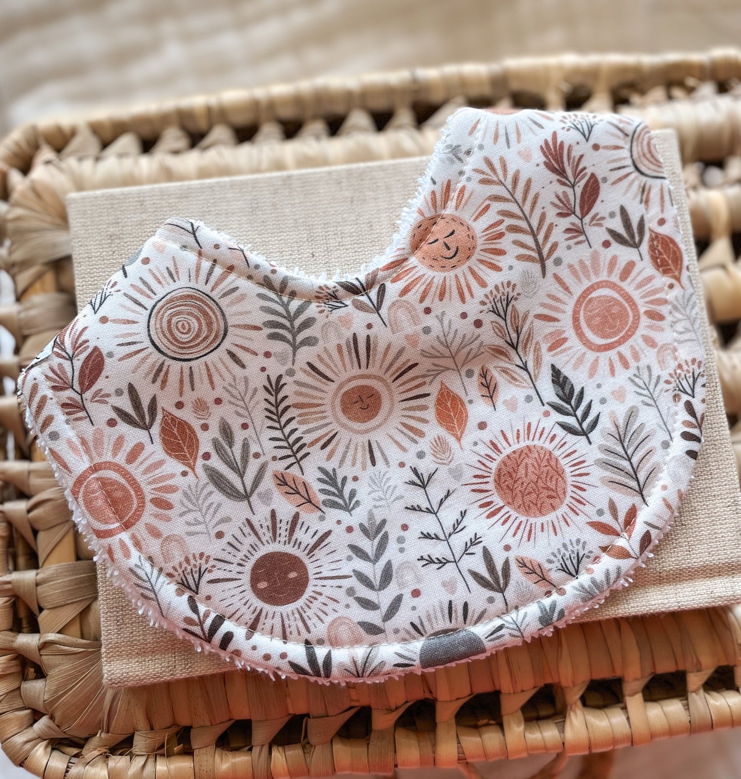Handmade baby bib with smiling sun faces, boho leaves, and earthy foliage in terracotta and grey tones; backed with absorbent terry towelling, ideal for newborns and teething babies.