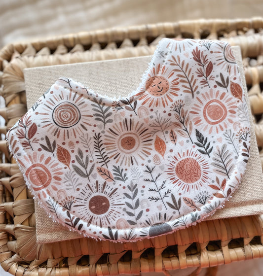 Handmade baby bib with smiling sun faces, boho leaves, and earthy foliage in terracotta and grey tones; backed with absorbent terry towelling, ideal for newborns and teething babies.