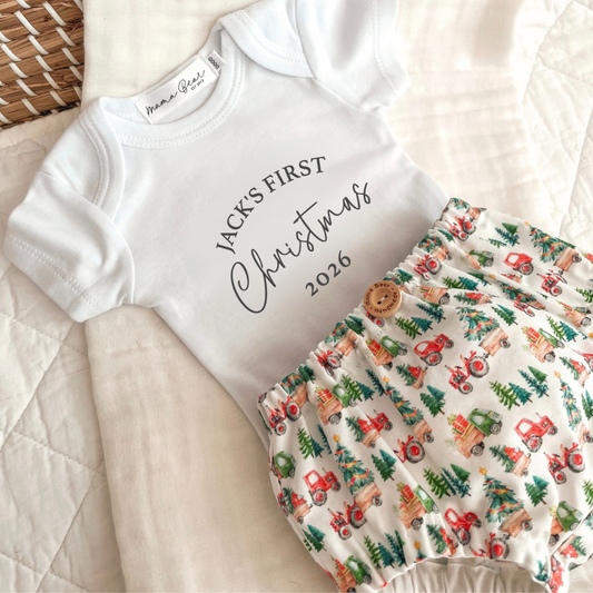 Personalised white baby onesie with 'Jack's first Christmas 2026'. Paired with some handmade nappy pants, fabric has red tractors and green trucks carrying Christmas presents and pine trees.