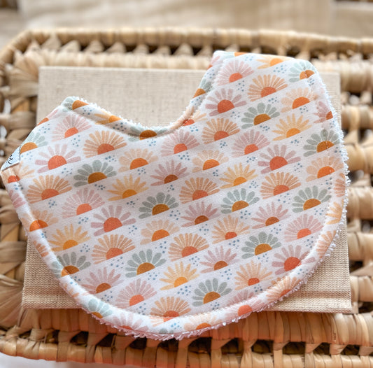 Handmade baby bib featuring a repeating floral sunrise pattern with daisy-like shapes in warm tones of blush, peach, mustard, and sage. The curved bib is made from soft cotton with a terry towelling backing, displayed on a neutral cloth and woven basket.