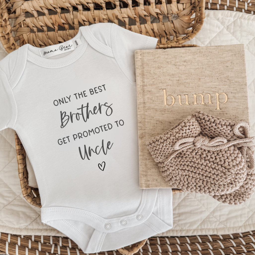 Pregnancy announcement flyaway. White baby bodysuit saying only the best brothers get promoted to Uncle. Laying next to a beige linen pregnancy journal and some knitted booties.