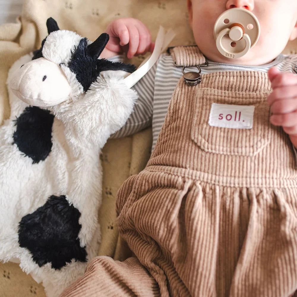Baby in brown overalls holding a plush cow toy and a  dummy.