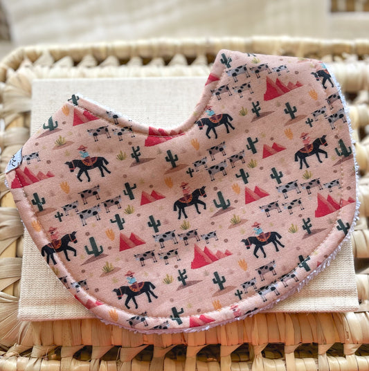 Handmade baby bib with cowboys riding horses, black and white cows, cacti, and desert hills on a dusty pink background; absorbent terry towelling backing, ideal for newborns and teething babies.