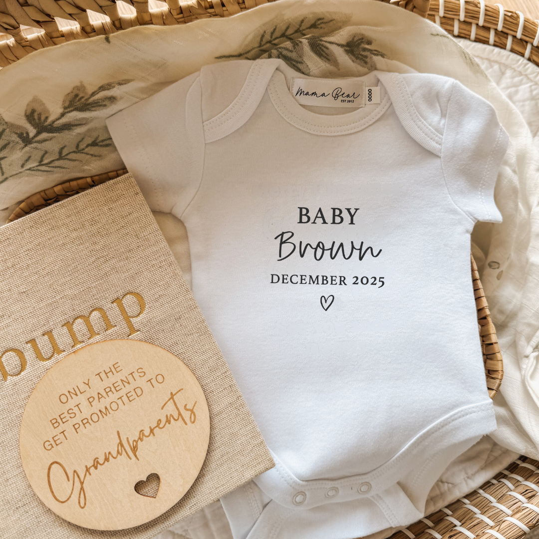 Flat lay of a white personalised baby onesie with the text “Baby Brown December 2025” in black serif and script fonts, finished with a small heart. Styled inside a wicker basket with a leafy muslin wrap, a beige “bump” pregnancy journal, and a wooden plaque that says “Only the best parents get promoted to grandparents.” A timeless and custom baby announcement onesie, perfect for pregnancy reveal photos, gift boxes, or keepsakes.