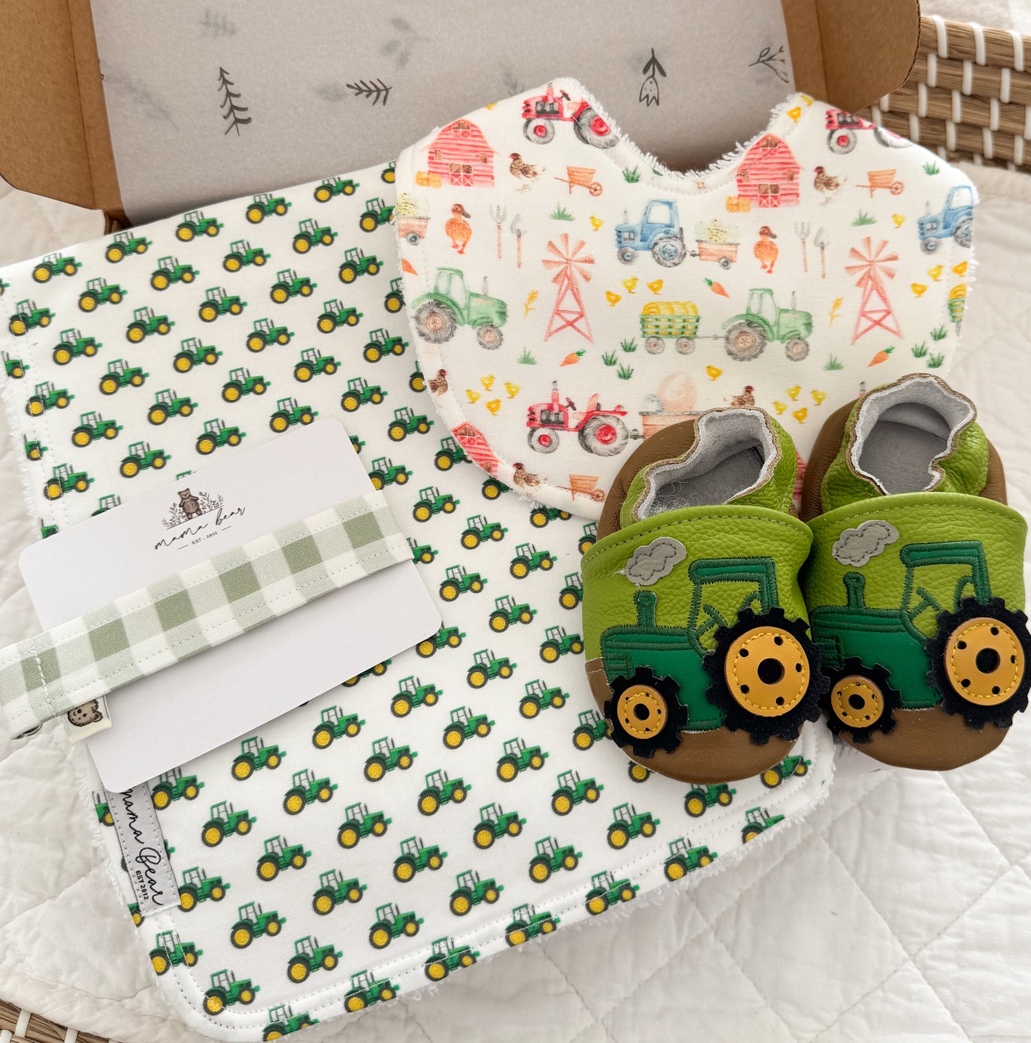 A baby gift set featuring a green tractor patterned burp cloth, a matching bib, a green and white gingham dummy clip, and a pair of green tractor theme baby shoes.