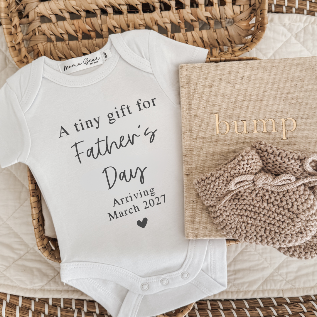 A white short-sleeve baby bodysuit printed with 'A tiny gift for Father's Day Arriving March 2027' in black script, styled in a wicker basket with a beige baby booties, a pregnancy journal labeled 'bump'.