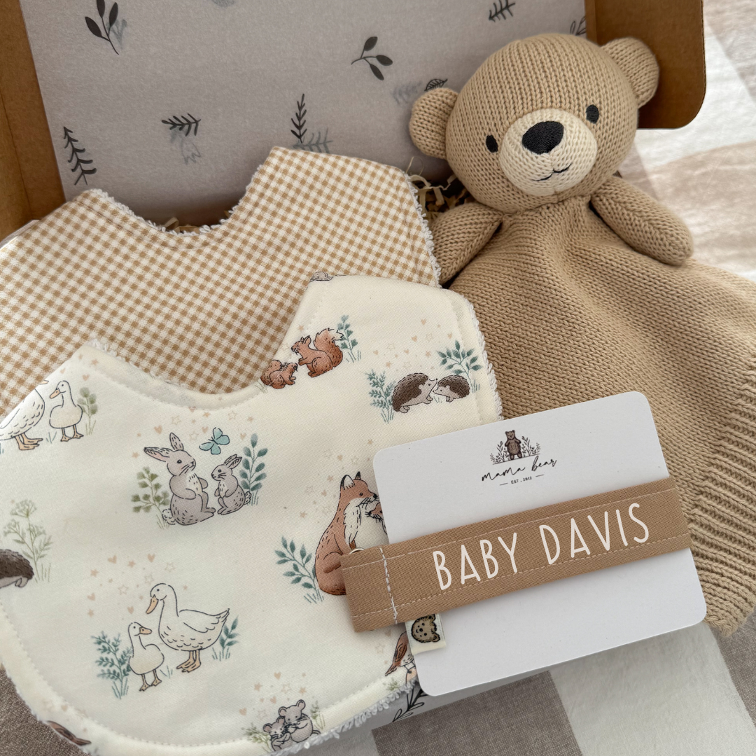 Baby shower gift idea, kraft gift box filled with 2 handmade baby bibs, one is a light brown mini gingham check, the other bib is an off white colour with illustrations of woodland creatures such as squirrels, rabbit, ducks, fox, hedgehog and mice. There is also a light brown teddy bear comforter and a light brown dummy clip with white text personalised with baby davis.