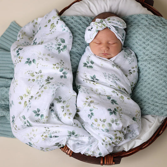 Newborn baby swaddled in a green leaf blanket with a matching headband, lying on a textured surface.