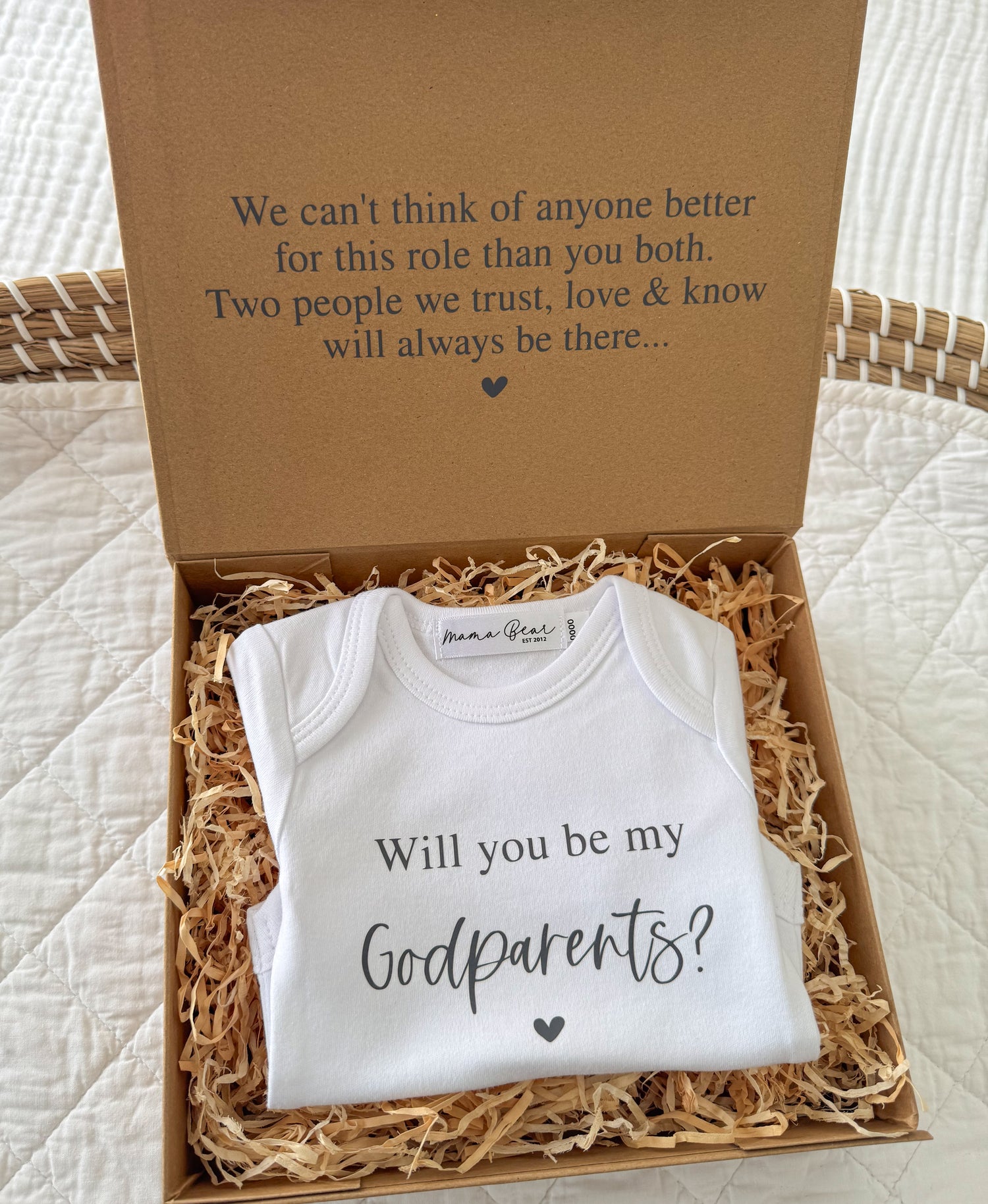 A kraft brown gift box with 'We can't think of anyone better for this role than you both. Two people we trust, love & know will always be there..." on the lid and inside a onesie that says 'Will you be my Godparents?' 