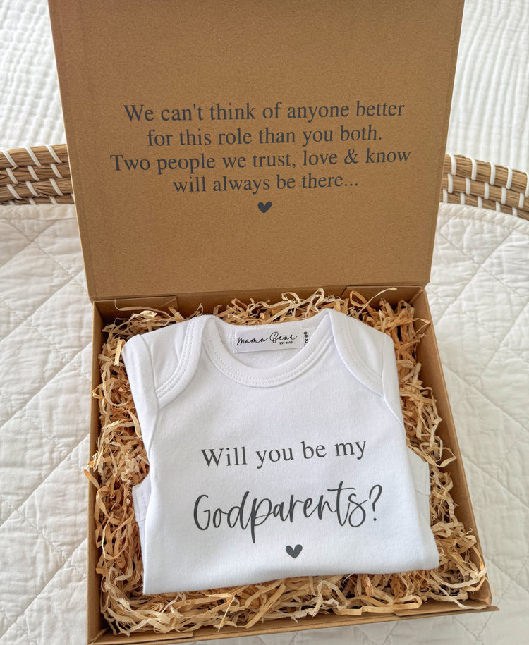 A kraft brown gift box with 'We can't think of anyone better for this role than you both. Two people we trust, love & know will always be there..." on the lid and inside a onesie that says 'Will you be my Godparents?' 