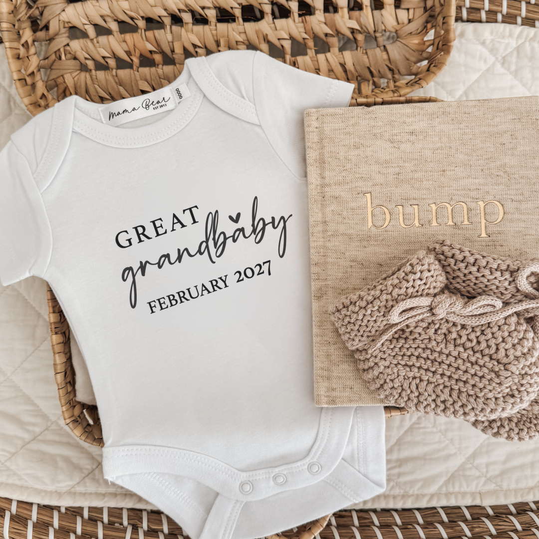 A white baby bodysuit with 'GREAT grandbaby FEBRUARY 2027' printed in black script, styled in a wicker basket with a beige photo book and a knitted beige booties.