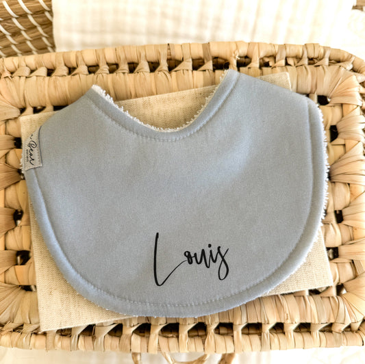 Handmade baby bib in a solid powder blue colour. Personalised with 'Louis' in script font black