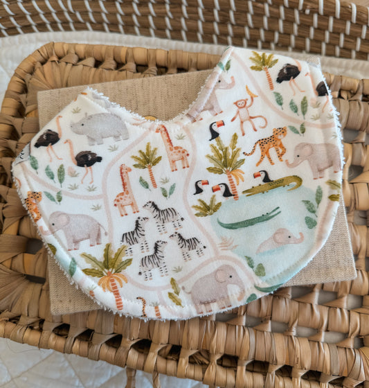A bib with a white background with mini illustrations of pastel wild jungle animals such as elephants, crocodiles, zebras, toucan birds, palm tree, zebras, water hole, monkey, cheatah, ostrich and rhinos  