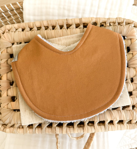 Handmade baby bib in a solid mustard, deep tan shade. Displayed on a wicker basket. 