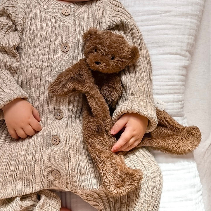 Child in a beige ribbed sweater holding a brown teddy bear on a white textured surface