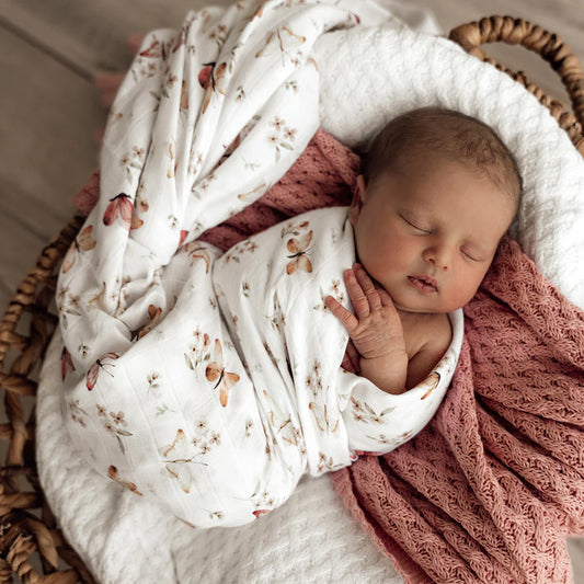 Newborn baby wrapped in a white blanket with floral & butterfly patterns, lying on a pink textured blanket inside a wicker basket.