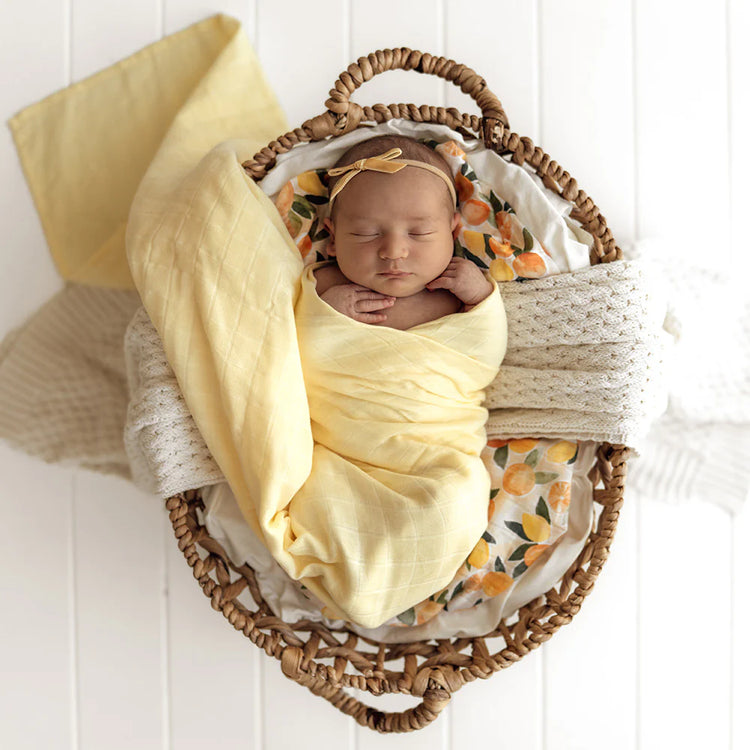 Newborn baby wrapped in a yellow blanket with floral headband in a woven basket on a white background