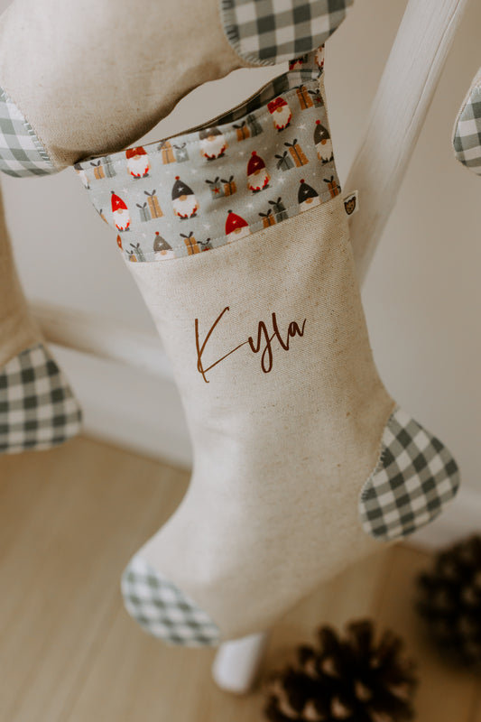 A personalised Christmas stocking with a gnome design, featuring the name 'Kylá' printed on the front, hanging on a mantel with pine cones decoration.
