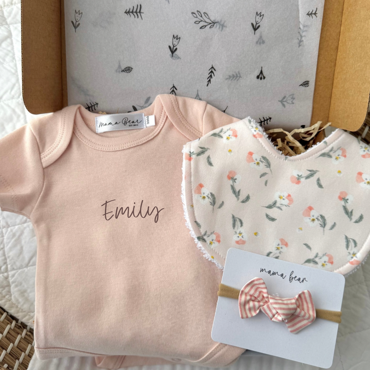 Baby Giftbox featuring a soft pink baby onesie with babies name in brown script vinyl on centre of onesie. A Soft blush baby bib with scattered illustrations of pink pansy flowers and a coordinating pink and white striped hair bow.  