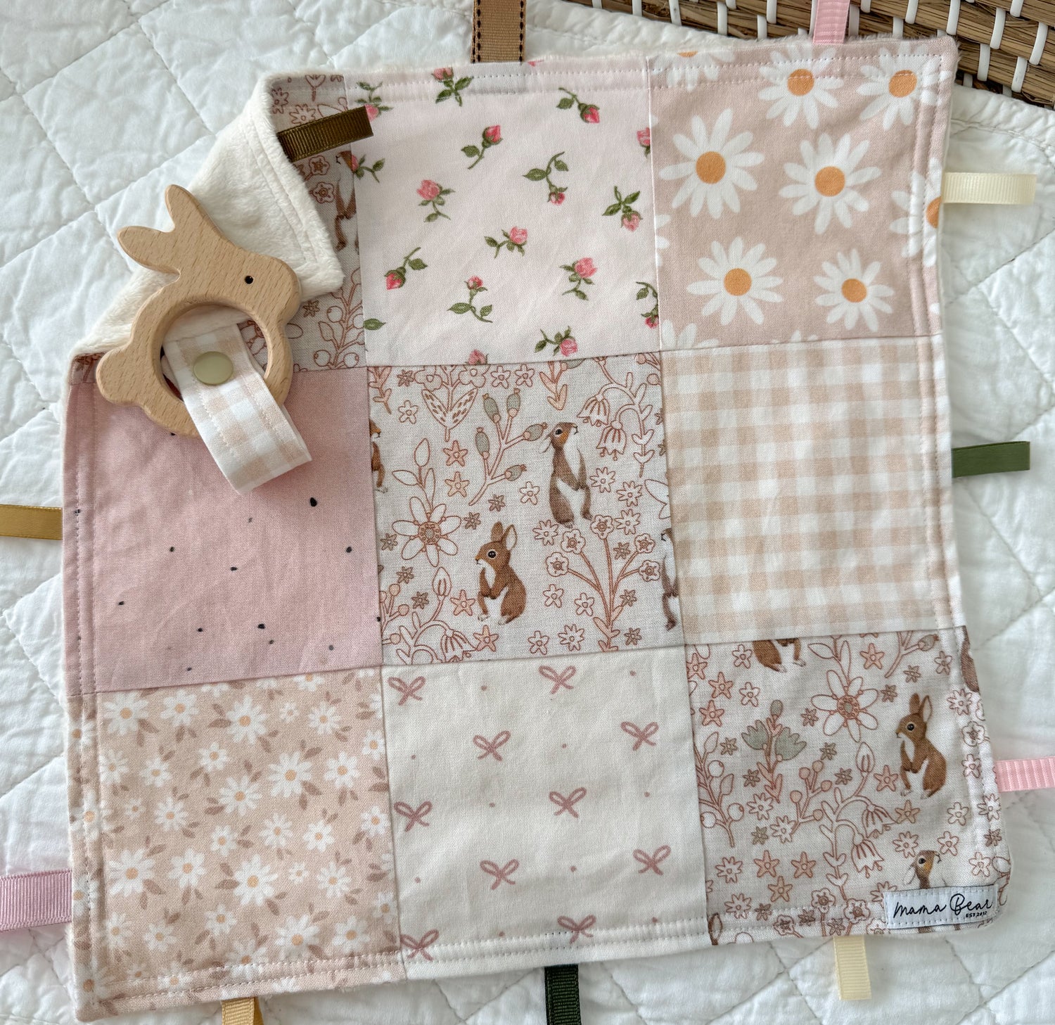 Patchwork baby blanket with floral and animal patterns, accompanied by a wooden teething ring on a white quilt.