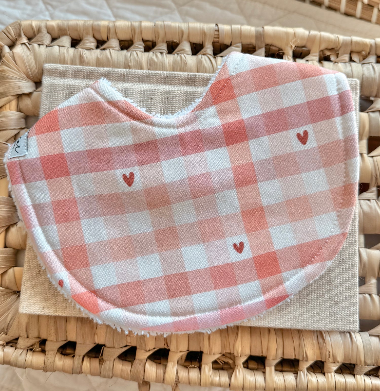 Flat lay of a handmade baby bib with a soft pink and white gingham print, scattered with tiny red hearts.