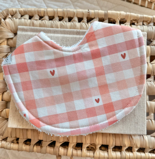 Flat lay of a handmade baby bib with a soft pink and white gingham print, scattered with tiny red hearts.