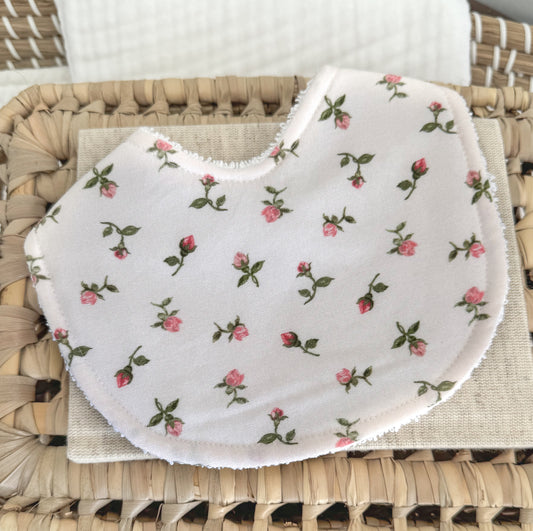 Baby girl bib in white with small blush pink rosebud print, styled on a woven basket.