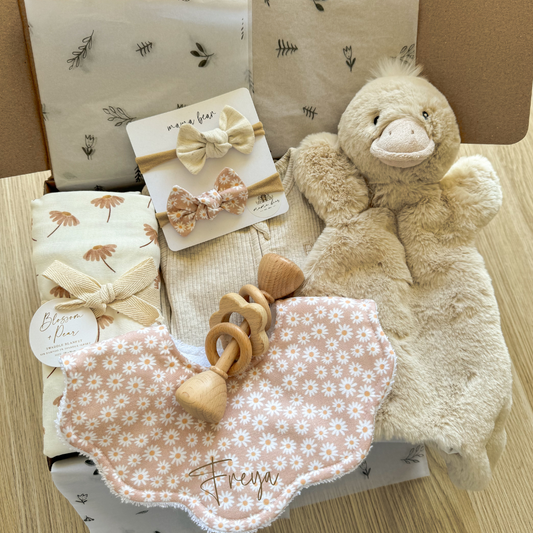 Personalised newborn baby girl gift. Box includes handmade baby bib with custom name on bib in brown script font, blush daisy swaddle, 2 matching baby hairbows, a soft duck toy, wooden flower rattle & a beige zip suit.  