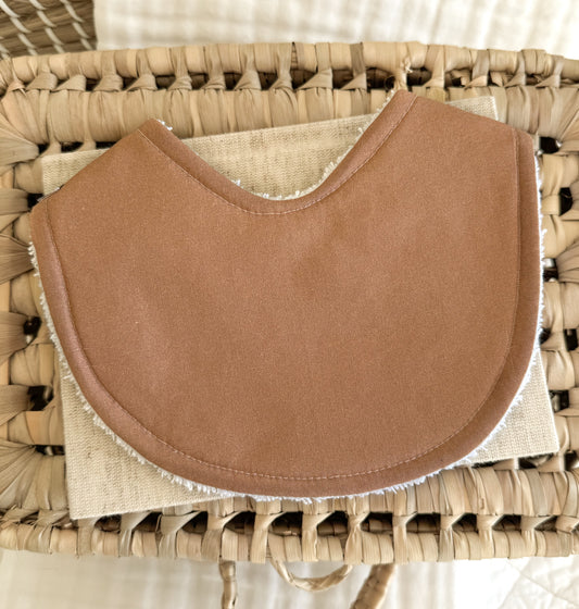 Flat lay of a handmade baby bib in a rich deep tan colour, displayed on a neutral linen cloth inside a woven basket.