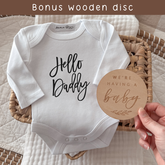 White baby onesie with 'Hello Daddy' text and a wooden disc reading 'We're having a baby' on a textured surface.