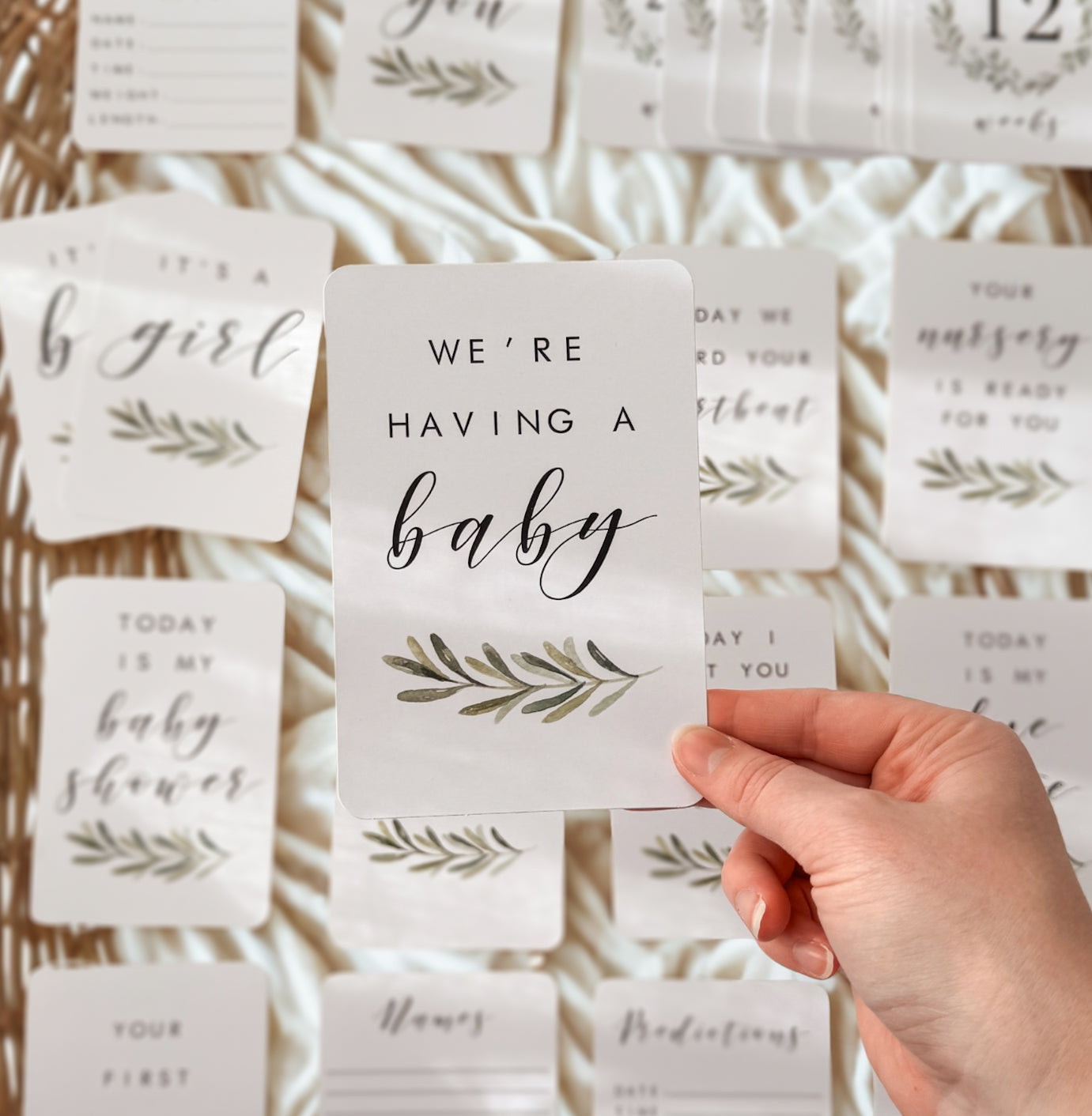 Hand holding a card with 'We're having a baby' text against a background of similar cards for pregnancy milestone cards.