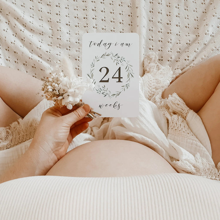 Pregnant belly with a card reading 'today i am 24 weeks' held by a hand against a textured white background.