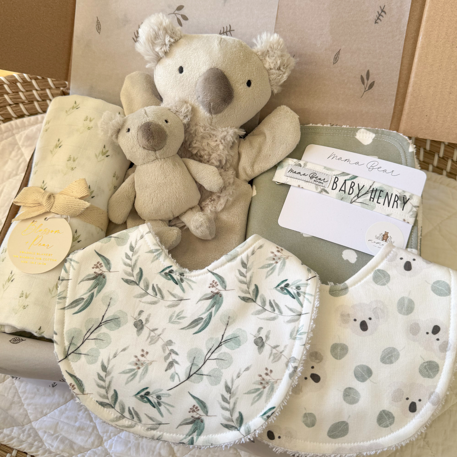 A baby gift hamper containing a plush koala toy, a smaller koala bear rattle, two bibs with a sage green leaf pattern, a handmade sage spot burp cloth, and a matching dummy clip personalised with baby henry.