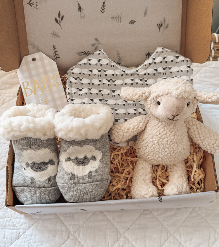 Unisex baby gift box featuring a sheep-themed bib, cozy lamb booties, and a soft plush lamb toy. The items are nestled in a gift box with shredded paper for a rustic, farm-inspired presentation—perfect for baby showers or newborn gifts.