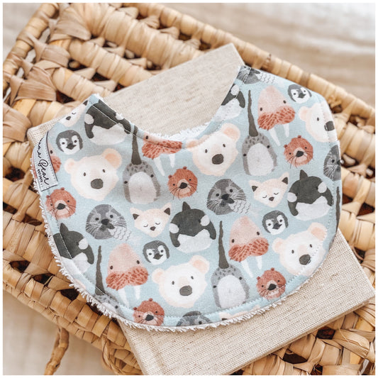 Baby bib with a light blue background featuring a variety of cute illustrated animal faces like polar bear, seal, penguins, killer whale styled on a woven basket and neutral linen cloth.