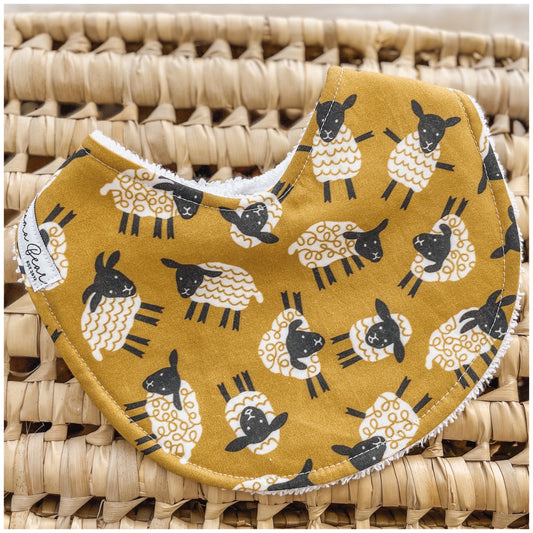 Baby bib with black and white sheep print on a mustard yellow fabric, styled on a woven basket.