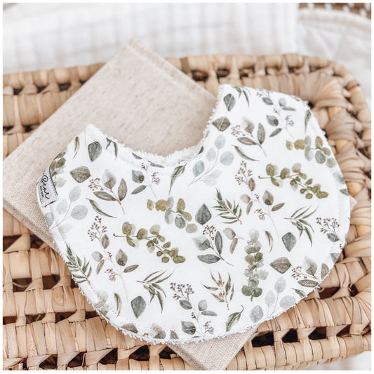 Handmade baby bib with a eucalyptus-themed print featuring green and grey leaves and sprigs on a white background. Bib is backed with beige terry towelling and styled on a woven basket.