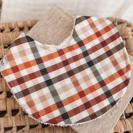 Baby bib with a checkered gingham pattern in earthy shades of rust, orange, olive green, and burgundy on a cream base. Backed with terry towelling for dribbles and spills.