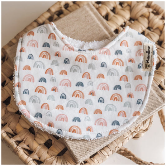 Handmade baby bib with a white base and small rainbow pattern in terracotta, blush, navy, and soft grey, backed with plush white terry towelling.
