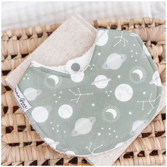 Sage green baby bib with a space-themed print including white planets, moons, constellations, and stars, displayed on a neutral linen cloth over a woven basket.