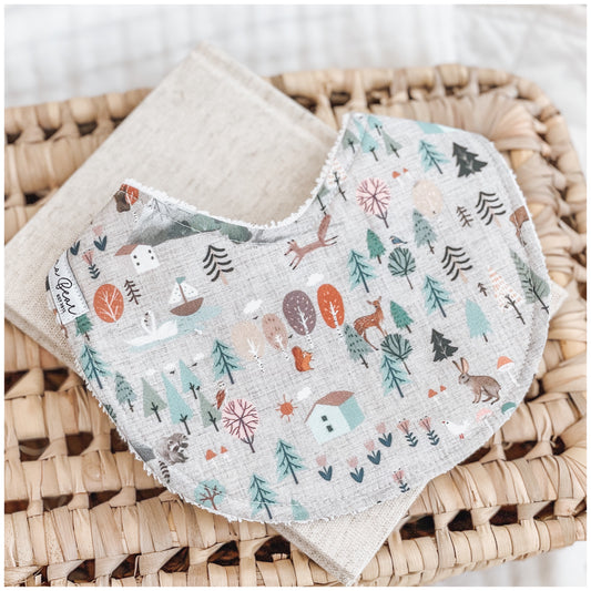 Handmade baby bib with a woodland village print featuring trees, cabins, deer, bunnies, and forest scenery in earthy and pastel colours on a light grey background. Bib is backed with beige terry towelling and displayed on a woven basket.